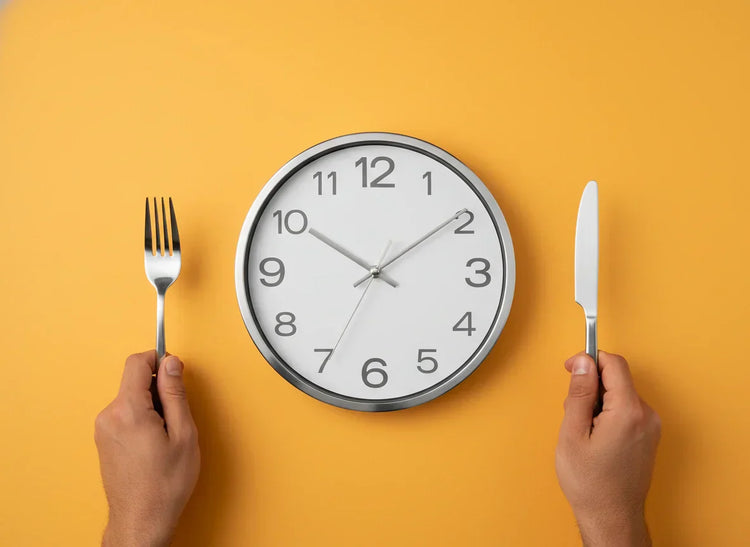 Modern round wall clock, white face, silver hands on marigold. Flat lay with hands holding silver dining fork & serrated knife.